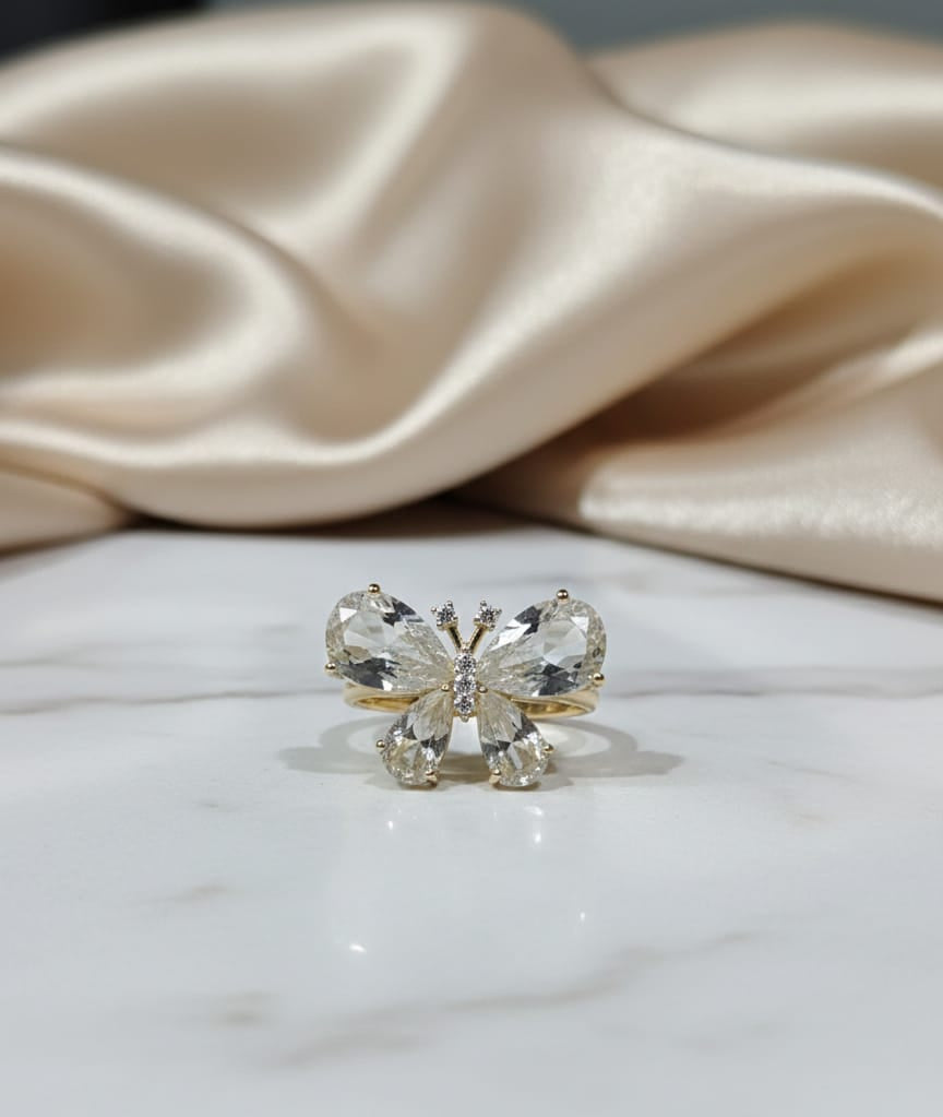 Butterfly-shaped ring with clear gemstones on a marble surface with beige fabric in the background