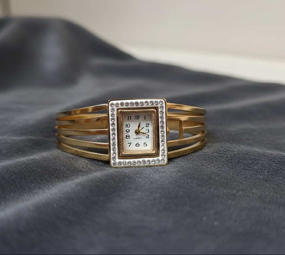 Gold bracelet with a square watch face on a dark fabric background